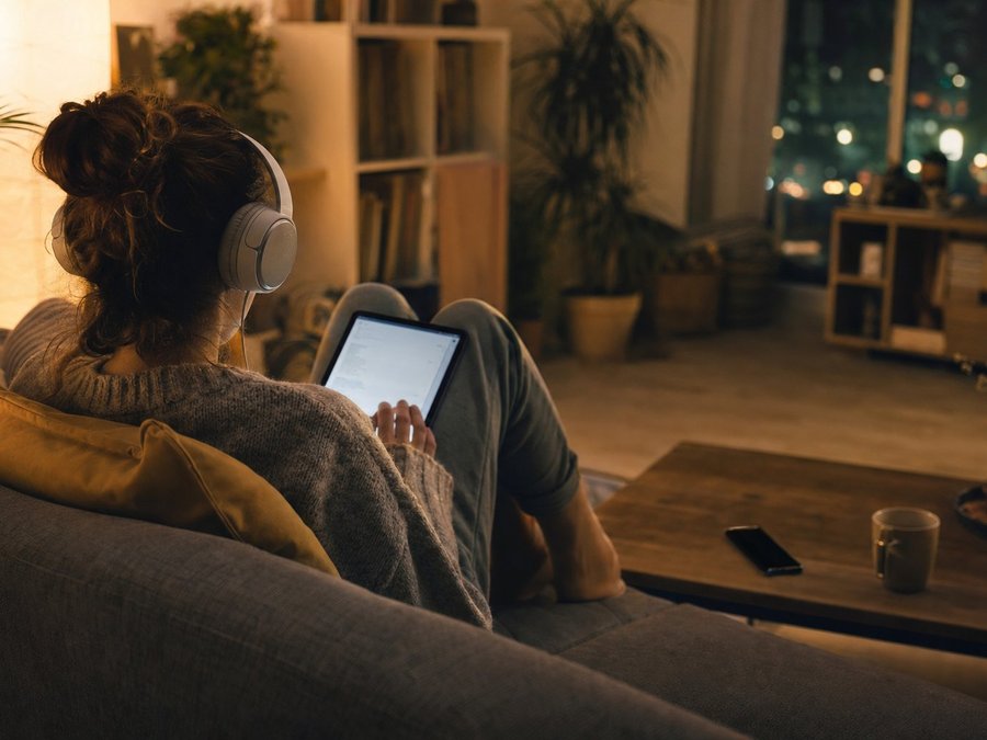 Person entspannt abends auf dem Sofa mit Tablet und Kopfhörern, warmes Wohnzimmerlicht, Smartphone auf dem Couchtisch